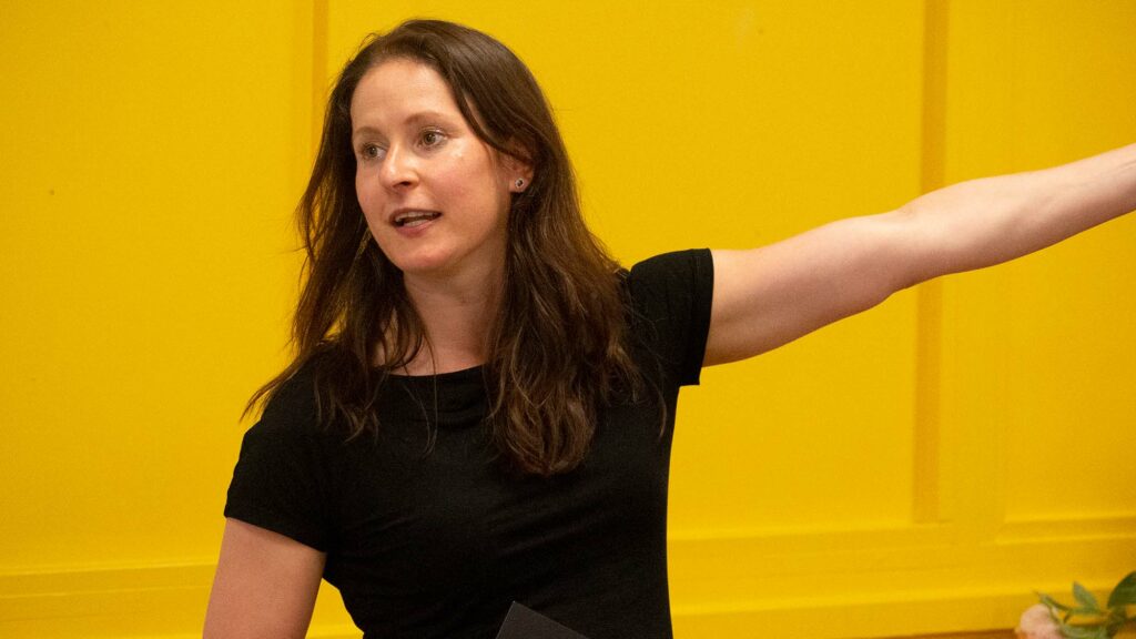 Ilona Eichhorn presenterend tegen gele achtergrond met armbeweging